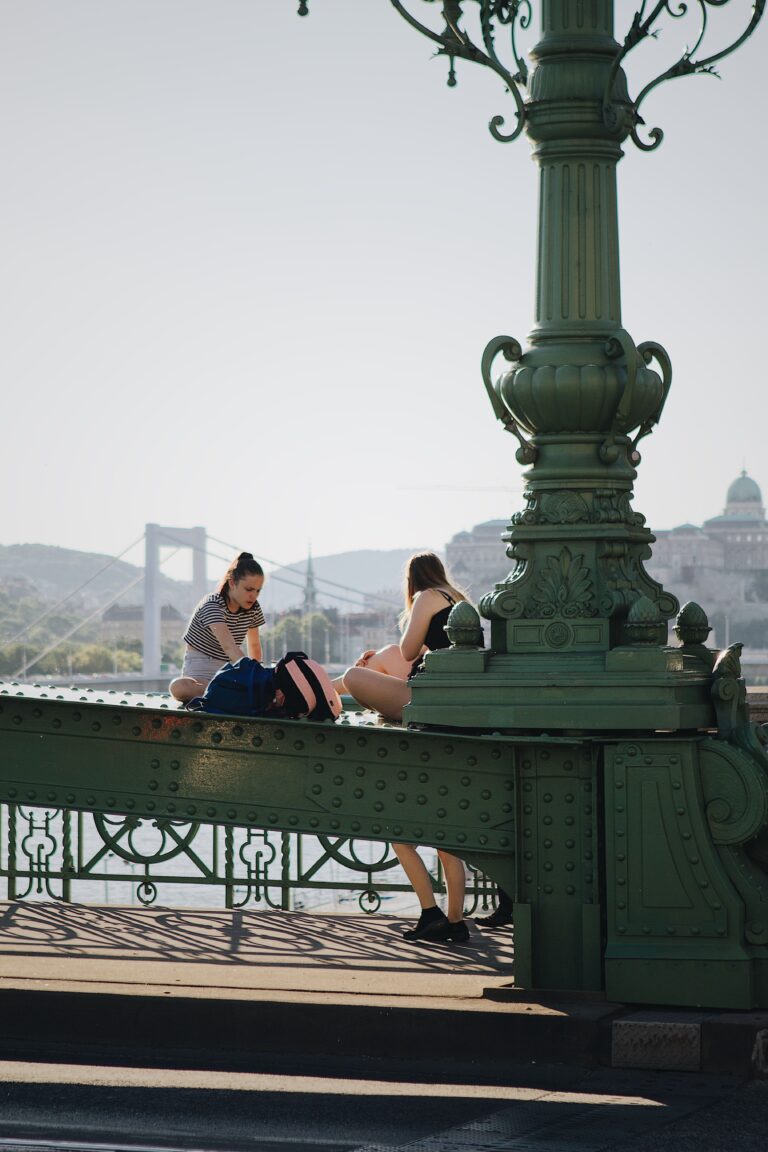 Two women sit on the Liberty Bridge in Budapest, Hungary, enjoying a sunny day with the cityscape in the background.